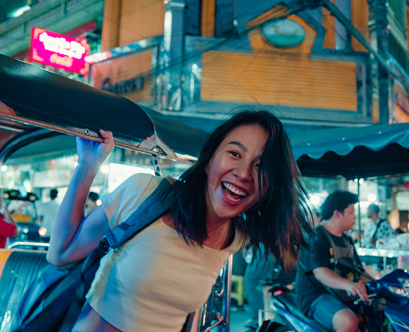 Eine junge Frau lacht und hält sich an einem Tuk-Tuk in einer belebten Stadt bei Nacht fest. Neonlichter und Gebäude sind im Hintergrund zu sehen, was auf eine lebhafte Atmosphäre hindeutet.