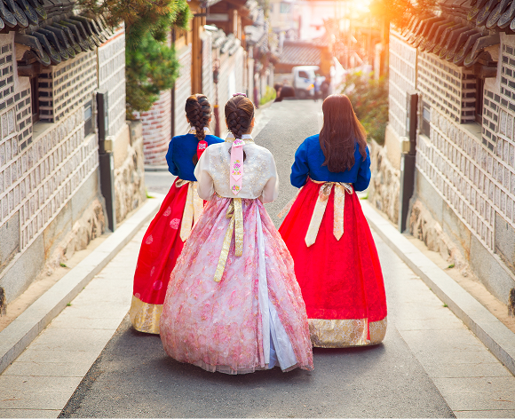 Drei Frauen in traditionellen koreanischen Hanbok-Kleidern gehen eine Gasse entlang. Die Kleider sind in leuchtenden Farben gehalten, mit weiten Röcken und verzierten Oberteilen. Die Szene spielt sich in einer historischen koreanischen Stadt ab, mit traditioneller Architektur im Hintergrund und Sonnenlicht, das durch die Gasse fällt.