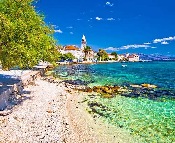 Ein malerischer Blick auf eine Küstenstadt mit kristallklarem türkisfarbenem Wasser, das sanft an einen Kiesstrand plätschert. Im Hintergrund sind traditionelle Gebäude mit roten Ziegeldächern und ein markanter Kirchturm zu sehen, die sich vor einem strahlend blauen Himmel mit vereinzelten Wolken abzeichnen. Die üppige grüne Vegetation am Ufer verleiht der Szene eine lebendige Note.