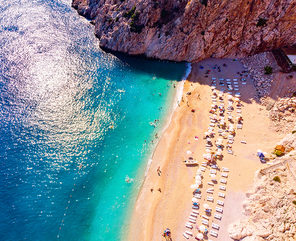Ein atemberaubender Blick auf eine sonnenverwöhnte Bucht mit türkisfarbenem Wasser, umgeben von steilen Felsklippen. Der Sandstrand ist mit Liegestühlen und Sonnenschirmen geschmückt und lädt zum Entspannen ein. Einige Urlauber schwimmen im kristallklaren Meer, während andere die Sonne auf dem Strand genießen.