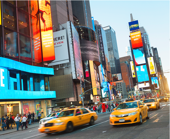 Eine belebte Straßenzene in New York City, mit vielen gelben Taxis, die auf der Straße fahren. Hohe Gebäude mit leuchtenden Werbetafeln säumen die Straße und schaffen eine energiegeladene städtische Atmosphäre. Menschen gehen auf den Bürgersteigen und tragen zum geschäftigen Treiben der Szene bei.